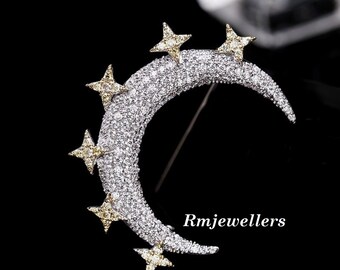 May include: A silver crescent moon brooch with a sparkling, clear crystal design. Five yellow stars with clear crystal accents are set around the moon. The brooch is accented with the text "Rmjewellers".