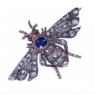May include: A detailed bee-shaped brooch featuring a central blue gemstone and many small clear stones. The wings and body are intricately designed, with ruby accents near the head. The brooch is set in a gold-toned metal.
