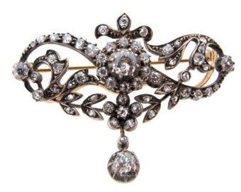 May include: A silver-toned brooch featuring a central cluster of clear stones surrounded by intricate scrollwork and leaf motifs. The piece is embellished with numerous small clear stones and includes a single dangling stone drop at the bottom.