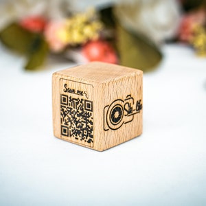 May include: A small wooden cube with a black and white QR code and the text "Scan me" on one side. The other side has a black and white camera icon with the text "Sle-Pics" on it.