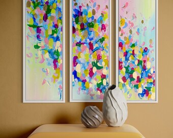 May include: Three abstract paintings in white frames with a colorful, abstract design of blue, green, pink, yellow, and gold. The paintings are hanging on a brown wall above a yellow dresser with two drawers.