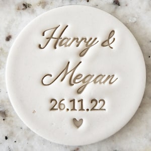 May include: White round ceramic tile with the text "Harry & Megan 26.11.22" and a small heart.