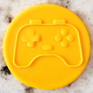 May include: A yellow plastic cookie cutter shaped like a video game controller. The controller has a plus sign, four round buttons, and two shoulder buttons.
