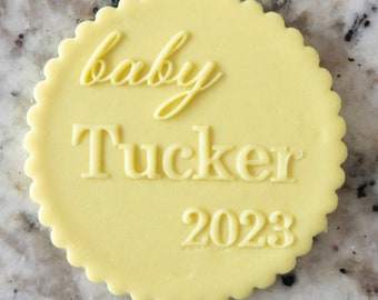 Könnte beinhalten: Ein runder gelber Tortendekor mit dem Text "baby Tucker 2023" in weiß.