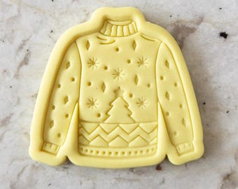 May include: A yellow cookie cutter in the shape of a holiday sweater. The sweater design includes a Christmas tree, snowflakes, and diamond shapes. The cookie cutter has a ribbed collar, cuffs, and hem.