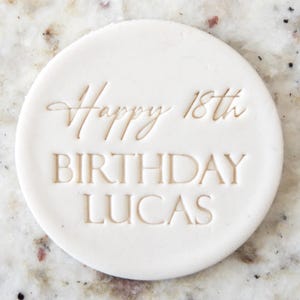 Puede incluir: Un adorno blanco y redondo para pastel con las palabras "Happy 18th Birthday Lucas" en relieve en una fuente simple y elegante. El texto está centrado en el adorno, que tiene una superficie lisa.