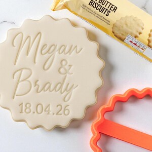 以下が含まれることがあります： 「Megan & Brady」と日付「18.04.26」が刻印された、明るい色のクッキー。オレンジ色のクッキーカッターとバタービスケットのパッケージも写っています。