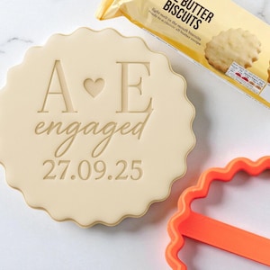 May include: A beige cookie cutter with the text "A ❤️ E engaged 27.09.25". The cookie cutter has a scalloped edge. A packet of butter biscuits is in the background, along with an orange cookie cutter.