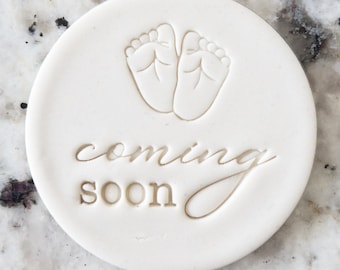 Puede incluir: Adorno redondo de cerámica blanca con una imagen de dos huellas de pies de bebé y el texto "coming soon" en una fuente cursiva.