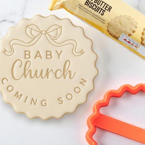 以下が含まれることがあります： 「BABY Church COMING SOON」の文字とリボン模様が入ったベージュ色のクッキー型。バタービスケットのパッケージとオレンジ色のクッキーカッターが背景にあります。