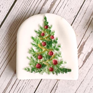 Puede incluir: Una galleta de jengibre blanca decorada con un árbol de Navidad verde con adornos rojos y una cinta dorada.