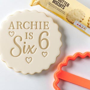 May include: A round, off-white cookie with the words "ARCHIE IS Six" and heart symbols. A package of butter biscuits is in the background. An orange, scalloped cookie cutter is in the foreground.