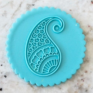 May include: A light blue paisley design stamp with a scalloped edge. The paisley pattern is detailed with swirls and floral elements. The stamp is likely used for crafts or baking.
