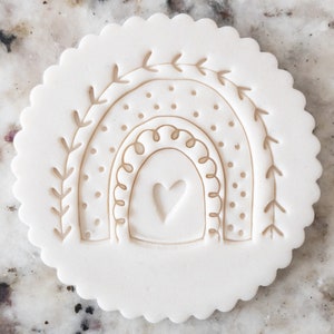 May include: White clay cookie cutter with a raised design of a rainbow with a heart in the center. The rainbow has a dotted pattern and a leafy design on the sides.