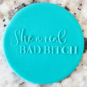 May include: A circular, turquoise-coloured object featuring the embossed phrase "She a real BAD BITCH" in white lettering. The object is set against a speckled, light-coloured surface.