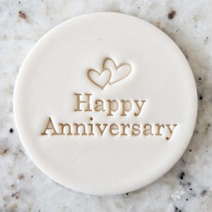 以下が含まれることがあります： 「Happy Anniversary」の文字と重なり合った2つのハートが描かれた白い丸いセラミックディスク