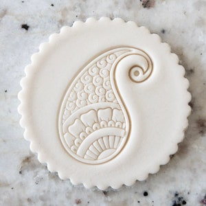May include: A white, round, scalloped-edged cookie cutter with a paisley design. The cutter is made of plastic and is approximately 5 cm in diameter.
