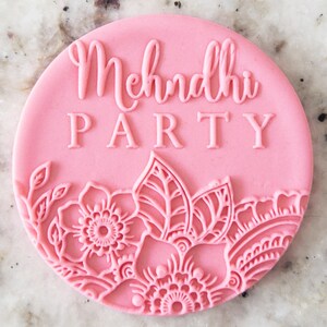 May include: Pink round cake topper with the text "Mehndi Party" in white lettering and a decorative floral design around the edge.