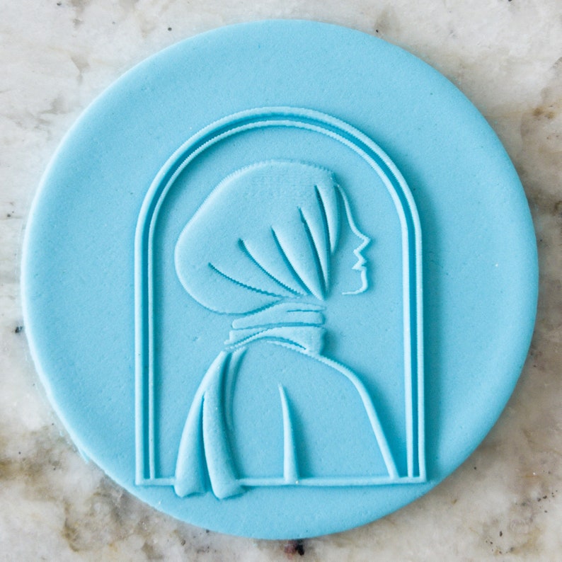 May include: A blue silicone cookie cutter in the shape of a woman wearing a headscarf. The cookie cutter is in a window frame design.