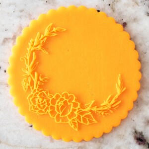 Peut inclure: Un biscuit rond de couleur jaune vif avec un bord festonné et un motif floral en relief sur la surface. Le motif représente une vigne avec des feuilles et des fleurs.