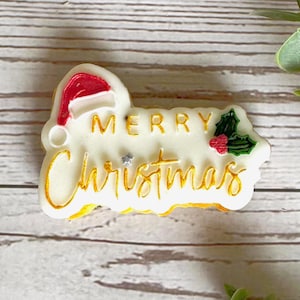 Puede incluir: Adorno de cerámica blanca con las palabras "Merry Christmas" en letras doradas. Un gorro de Papá Noel rojo y una rama de acebo verde están en el adorno.