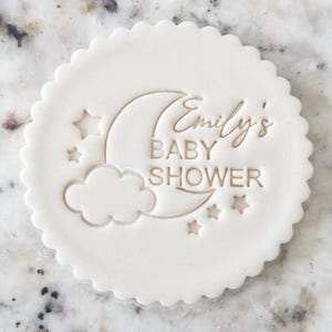 Puede incluir: Un cortador de galletas redondo, blanquecino, con borde festoneado. El diseño incluye una luna creciente, estrellas y una nube, con el texto "Emily's BABY SHOWER" en letra cursiva. El diseño está en relieve.