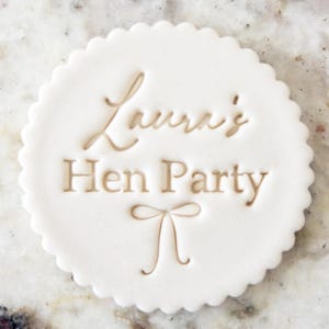 Puede incluir: Un fondant blanco y circular con borde festoneado. El texto "Laura's Hen Party" está grabado en color marrón claro, con un diseño de lazo debajo. El fondant está sobre una superficie clara.