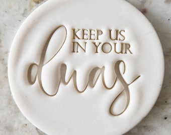 May include: White ceramic round stamp with the text "Keep us in your prayers" in a gold font.