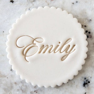 May include: White round cake topper with a scalloped edge and the name "Emily" embossed in gold.