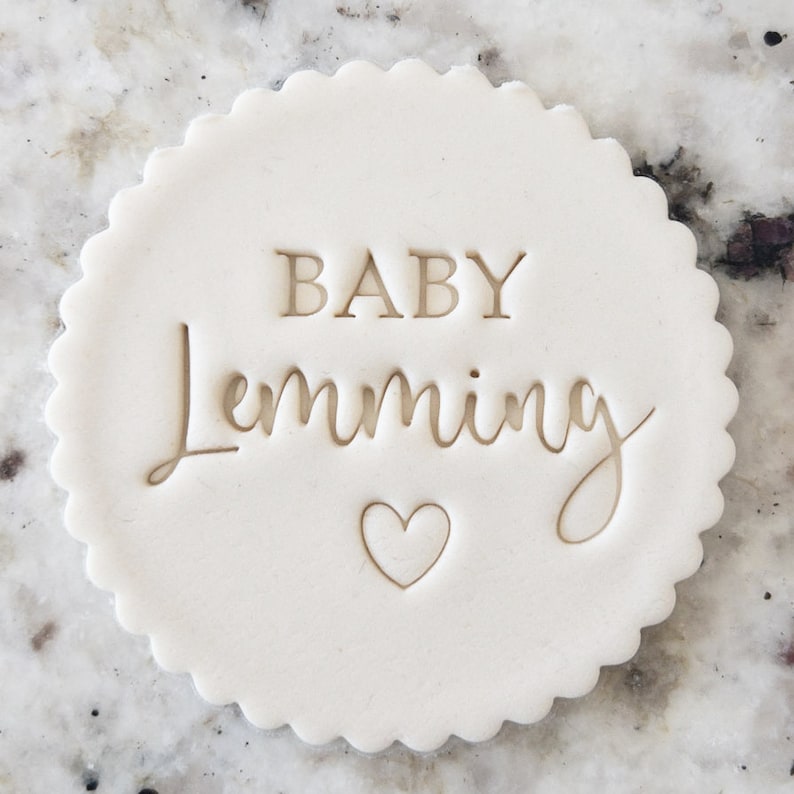 May include: White round cookie with the words "BABY Lemming" and a small heart in the center.