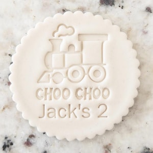 Op de afbeelding: Een ronde, gebroken witte koek met een geschulpte rand. De koek heeft een reliëf treindesign met de tekst "CHOO CHOO Jack's 2". De trein heeft een schoorsteen, wielen en een rechthoekig lichaam. Het ontwerp is gecentreerd.