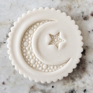 Pode incluir: Um cortador de biscoitos de argila branca com um design de lua crescente e estrela. A lua e a estrela são texturizadas com pequenos círculos.