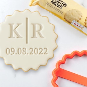 Puede incluir: Una galleta crema con borde festoneado con las iniciales "KR" y la fecha "09.08.2022" en relieve. Un paquete de galletas de mantequilla está en el fondo, junto con un cortador de galletas naranja.