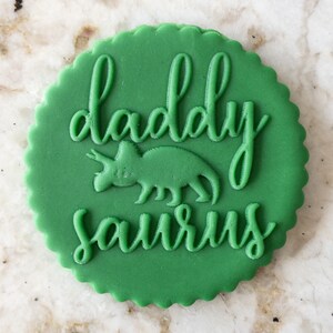 Puede incluir: Galletas de fondant verde con el texto "daddy saurus" en glaseado blanco y una silueta de dinosaurio en el centro.