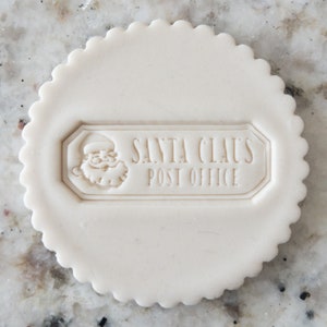 May include: White round cookie cutter with a scalloped edge. The cutter features a raised design of Santa Claus's head and the text "Santa Claus Post Office".