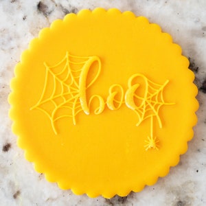 May include: A yellow round cookie decorated with a white spiderweb and the word "boo" in white icing.