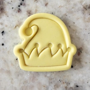 Puede incluir: Cortador de galletas de plástico amarillo con forma de gorro de elfo navideño con la parte superior puntiaguda y una corona en la parte inferior.