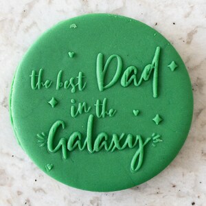Puede incluir: Cortador de galletas redondo verde con el texto "the best Dad in the Galaxy" en una fuente blanca de escritura.