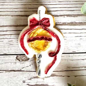 Puede incluir: Una galleta con forma de adorno navideño decorada con glaseado blanco, un lazo rojo y un adorno dorado. Cintas de glaseado rojo y detalles de cuentas plateadas completan el diseño festivo. La galleta está sobre una superficie de madera.