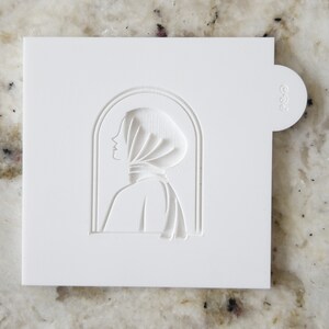 May include: White plastic stencil with a cutout design of a woman's profile wearing a headscarf. The design is framed by an arched window shape.