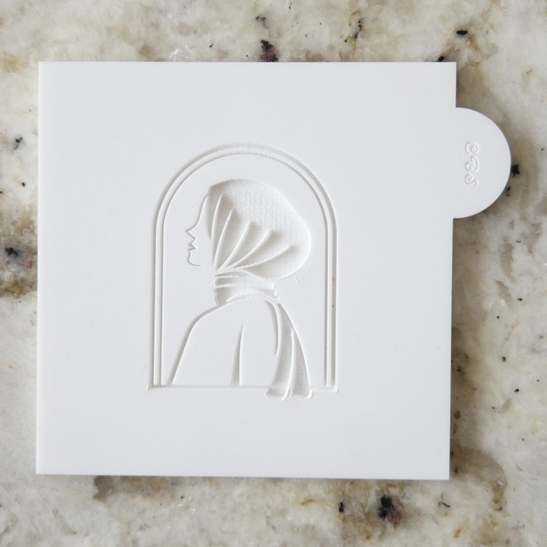 May include: White plastic stencil with a cutout design of a woman's profile wearing a headscarf. The design is framed by an arched window shape.