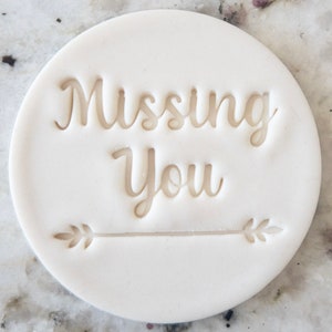 Op de afbeelding: Ronde witte koekjesuitsteker met de woorden "Missing You" en een pijl met bladeren aan beide kanten.