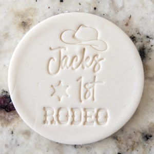 May include: A round, off-white disc with embossed text and a cowboy hat design. The text reads "Jacks 1st RODEO" in a mix of script and block lettering, with a star symbol. The disc is likely a decorative element for a rodeo-themed event.