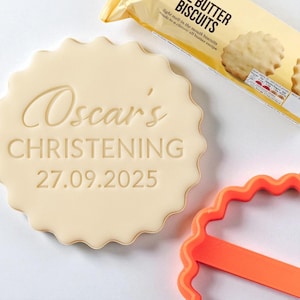 Puede incluir: Una galleta clara con borde festoneado con la inscripción "Oscar's CHRISTENING 27.09.2025" se muestra. Un paquete de galletas de mantequilla y un cortador de galletas naranja también son visibles.