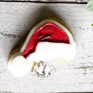 Puede incluir: Una galleta de azúcar blanca y roja con forma de gorro de Papá Noel. La galleta está decorada con glaseado rojo y blanco.