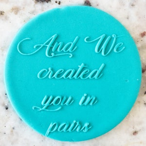 Op de afbeelding: Een ronde turquoise blauwe stempel met de tekst "And We created you in pairs" in een cursief schrift.