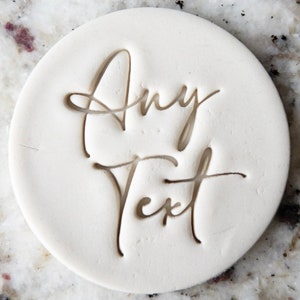 May include: White round clay stamp with the words "Any Text" engraved in a cursive font.