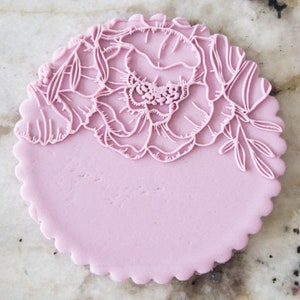 Può includere: Un topper per torta in pasta di zucchero rosa con un delicato design floreale. Il design presenta un grande fiore con dettagli intricati e foglie più piccole.