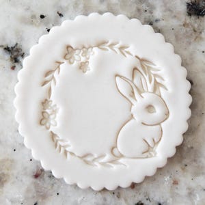 May include: White ceramic cookie cutter with a detailed design of a rabbit sitting in a wreath of leaves and flowers.