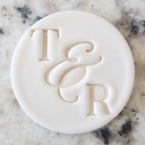 Peut inclure: Un motif rond blanc en relief avec les lettres "T", "&" et "R". Les lettres sont dans une police serif classique et légèrement en relief. Le motif est centré sur un fond blanc, idéal pour la papeterie de mariage.